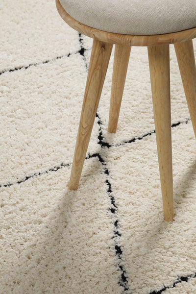 Hocker mit Holzbeinen und gepolsterter Sitzfläche auf einem Hochflor-Teppich mit geometrischem Muster.