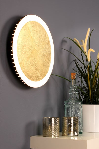 Runde LED-Wandleuchte mit Blattgold-Optik und hellem Lichtring an einer grauen Wand über einer weißen Ablage mit Dekoration.