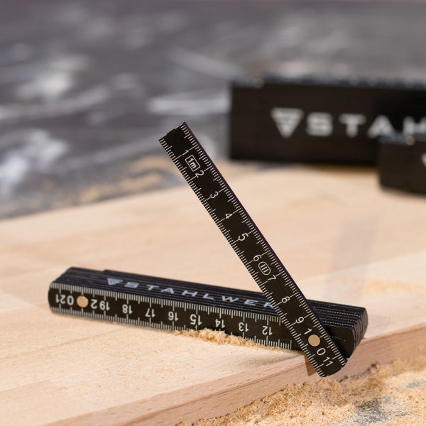 Ein Meter langer schwarzer Gliedermaßstab von Stahlwerk auf einem Holzbrett mit Sägespänen.
