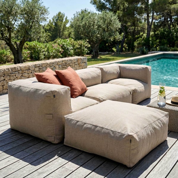 Modulares Outdoor-Lounge-Set aus Stoff mit Sofa und Hocker auf einer Holzterrasse am Pool, umgeben von Bäumen und einer Natursteinmauer.
