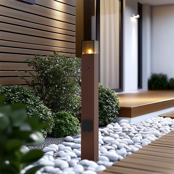 Moderne Wegeleuchte in Rostoptik mit Glaseinsatz in einem Garten mit weißen Kieselsteinen und grünen Sträuchern vor einer Holzfassade.
