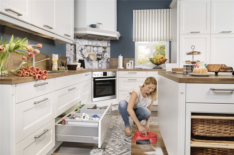Helle, organisierte Küche mit weißen Schränken und Holz-Arbeitsplatten. Eine Frau fegt den Boden mit Handfeger und Kehrschaufel.