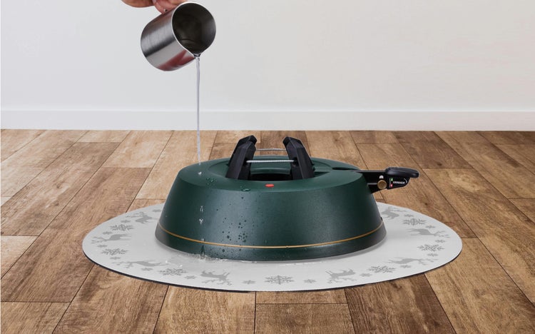 Wasser wird in einen dunkelgrünen Christbaumständer gegossen, der auf einer runden Unterlagsmatte mit winterlichem Muster auf einem Holzboden steht.