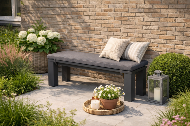Dunkelgraue Gartenbank aus Metall mit Sitzkissen und Dekokissen auf einer Terrasse vor einer Mauer, dekoriert mit Pflanzen, Laterne und Kerzen.