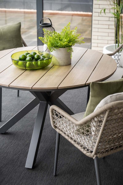 Runder Gartentisch mit Holzlatten und Metallgestell neben geflochtenen Gartenstühlen mit Kissen auf einer Terrasse.