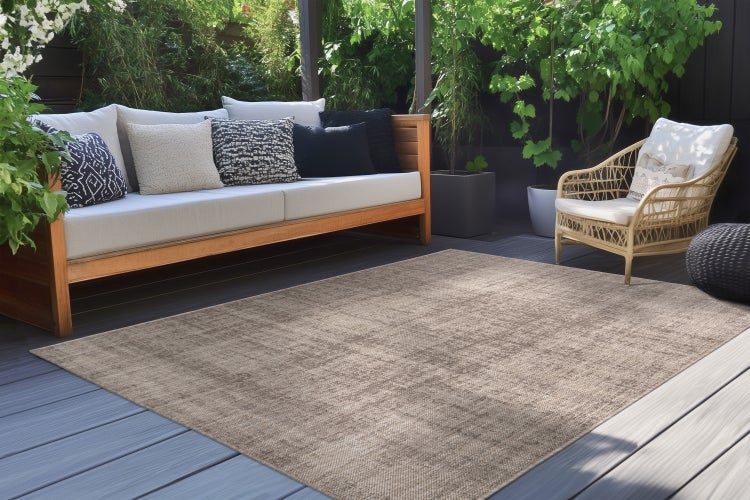 Ein moderner, rechteckiger Außenteppich auf einer Holzterrasse mit einem Loungesofa und einem Rattansessel in einem gemütlichen Garten.