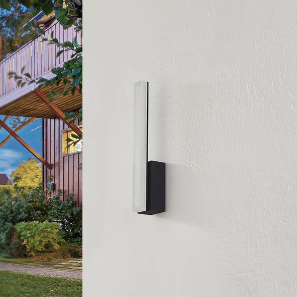 Moderne vertikale LED Außenwandleuchte an einer weißen Wand im Außenbereich mit Garten und Holzhaus im Hintergrund.