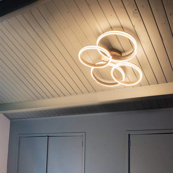 Moderne LED-Deckenleuchte mit vier ineinandergreifenden Ringen, montiert an einer Paneeldecke aus Holz.