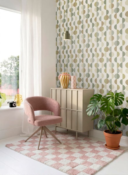 Modernes Zimmer mit geometrischer Vliestapete in Grün und Beige, rosa Sessel, kariertem Teppich, Sideboard und einer Monstera-Pflanze.