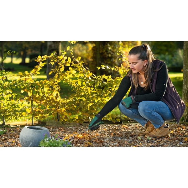 Eine Frau hockt im Garten und entfernt Laub mit einem Laubgreifer.