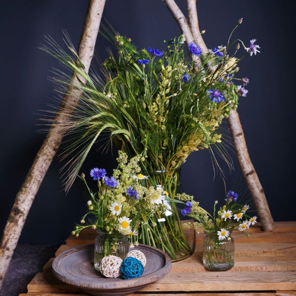 Rustikale Dekoration mit Wildblumen wie Kornblumen und Margeriten in Glasvasen und Birkenstämmen auf einer Holzpalette.