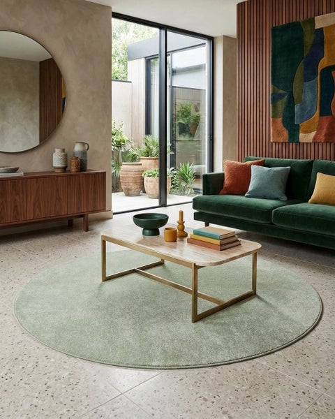 Moderner Wohnraum mit rundem grünem Teppich auf Terrazzoboden, Marmor-Couchtisch und grünem Samtsofa vor einer Holzwand.