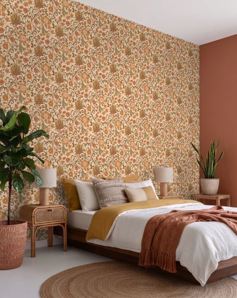 Schlafzimmer mit floral gemusterter Tapete, Holzbett mit weißer und senfgelber Bettwäsche, Nachttisch aus Rattan und Zimmerpflanzen.