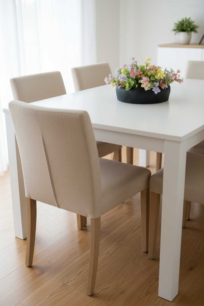 Moderner, heller Essbereich mit weißem Esstisch und beige gepolsterten Stühlen auf hellem Holzboden mit dekorativem Blumengesteck.