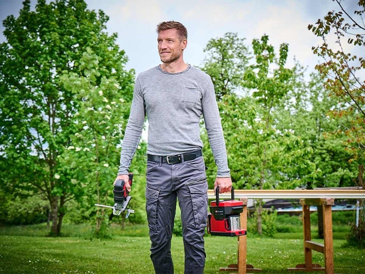 Ein Heimwerker im Garten hält eine Akku-Stichsäge und eine Akku-Kaffeemaschine, im Hintergrund steht eine Werkbank aus Holz.