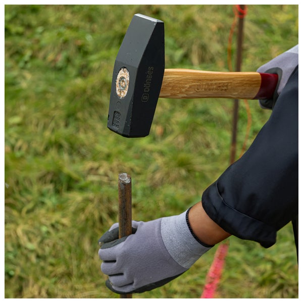 Eine Person schlägt mit einem Dönges Hammer einen Metallstab in den Boden und trägt dabei graue Arbeitshandschuhe.