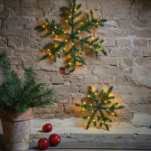 Zwei künstliche Tannenzweig-Schneeflocken mit Lichtern hängen an einer Ziegelwand, neben einem Topf mit Tannenzweigen und Äpfeln.