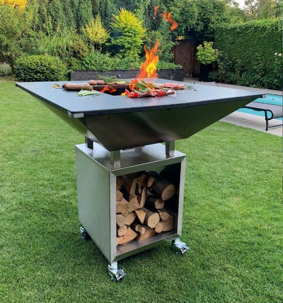 Mobiler Feuerstellengrill aus Metall mit Holzaufbewahrung, Grillplatte und offenem Feuer, im Garten auf Rasenfläche platziert.