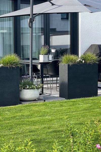 Moderne Terrasse mit großen schwarzen rechteckigen Pflanzkübeln, grauem Ampelschirm und Gartenmöbeln auf einem Holzdeck neben einem grünen Rasen.
