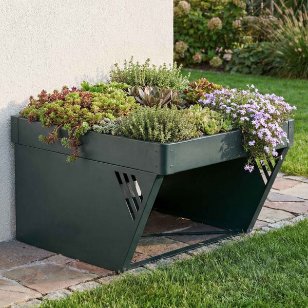Modernes grünes Hochbeet aus Metall mit integriertem Stauraum, bepflanzt mit Sukkulenten und lila Blumen, an einer Hauswand im hellen Garten.