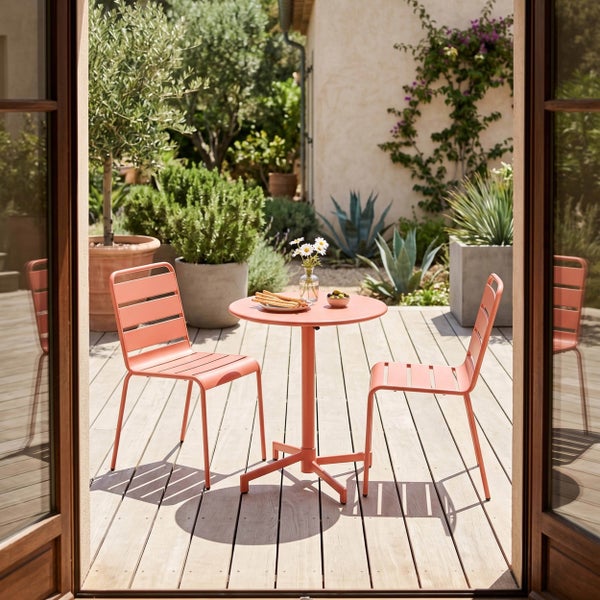Modernes Gartenmöbel-Set aus Metall mit rundem Tisch und zwei Stühlen auf einer Holzterrasse in einem hellen Garten mit Pflanzen.