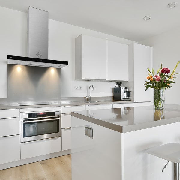 Moderne, helle Küche mit weißen Fronten, einer Kücheninsel mit grauer Arbeitsplatte, Dunstabzugshaube aus Edelstahl und Einbaubackofen.
