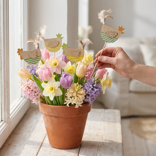 Hand steckt Holzstecker mit Huhnmotiv in Blumentopf mit Frühlingsblumen wie Tulpen, Narzissen und Hyazinthen auf Fensterbank zur Osterdekoration.