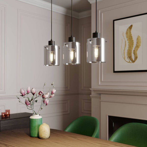 Dreiflammige Pendelleuchte mit zylindrischen Glasschirmen über einem Holzesstisch in einem hellen Raum mit Wandleisten und Kamin.