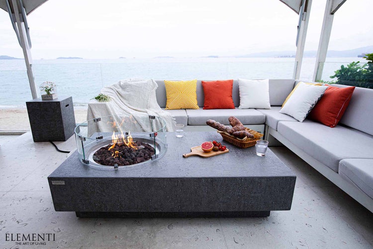 Lounge-Set auf Terrasse mit grauem Beton-Gaskamin-Tisch, Sicherheitsglas, L-Sofa mit Kissen und Meerblick.