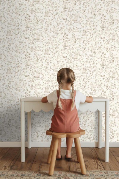 Kinderspielzimmer mit Vliestapete mit Hasenmotiven und Blumenranken, einem weißen Kinderschreibtisch und einem Holzhocker.