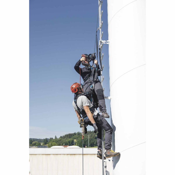 Zwei Personen mit Absturzsicherung und Sicherheitsgurten bei Höhenarbeiten an einer Leiter an einem Industrieturm.