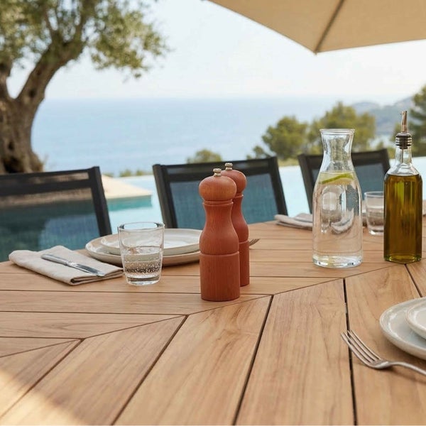 Gartentisch aus Holz mit Fischgrätenmuster, gedeckt mit Salz- und Pfeffermühlen, Karaffe und Gläsern auf einer Terrasse mit Pool und Meerblick.