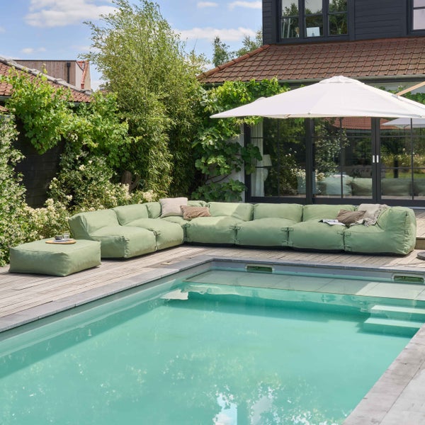 Modulares Outdoor-Lounge-Sofa in Olivgrün auf einer Holzterrasse neben einem Pool mit Sonnenschirm und üppiger Bepflanzung.