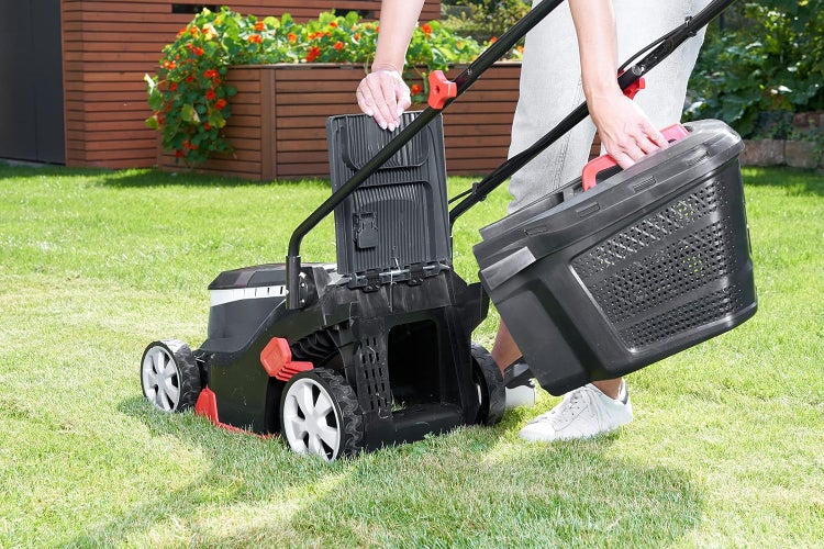 Helle Gartenszene mit einer Person, die einen Kunststoff-Fangkorb an einen schwarzen Elektrorasenmäher auf einer Rasenfläche im Garten anbringt.