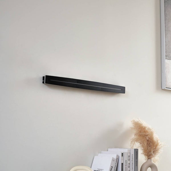 Moderne, schwarze, lineare Wandleuchte an einer hellen Wand in einem hellen Raum. Darunter Bücher und eine Vase mit Pampasgras.