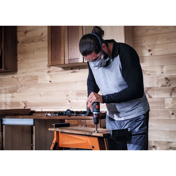 Mann bohrt mit Akkubohrmaschine ein Loch in ein Holzbrett, getragen von einer Werkbank, mit persönlicher Schutzausrüstung