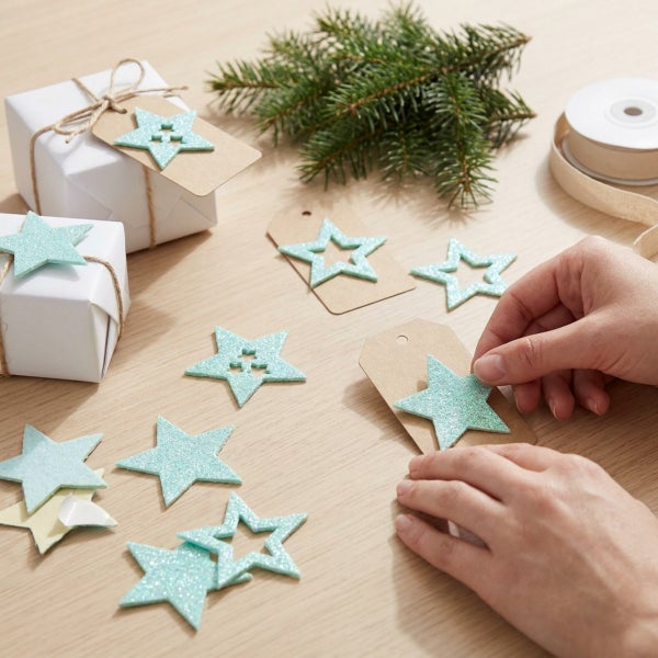 Hände beim Dekorieren von Geschenkanhängern mit glitzernden Sternaufklebern auf einem Holztisch mit Tannenzweigen.