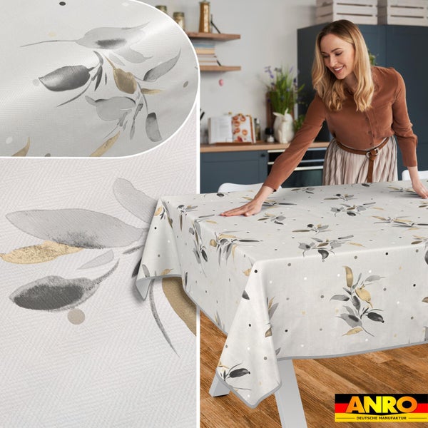Frau legt eine abwaschbare Tischdecke mit Blattmuster und Punkten auf einen Esstisch in einer hellen Küche. ANRO Deutsche Manufaktur Logo.
