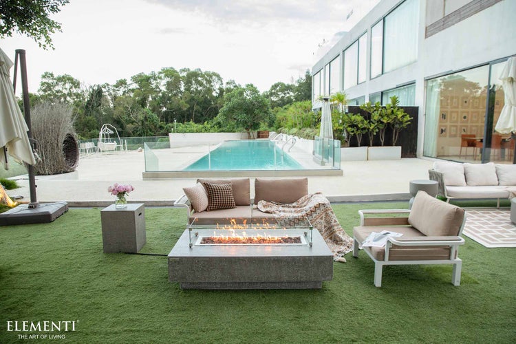 Moderner Außenbereich mit rechteckiger Gasfeuerstelle, weißer Gartenlounge mit beigen Polstern, Pool im Hintergrund und Elementi Logo.