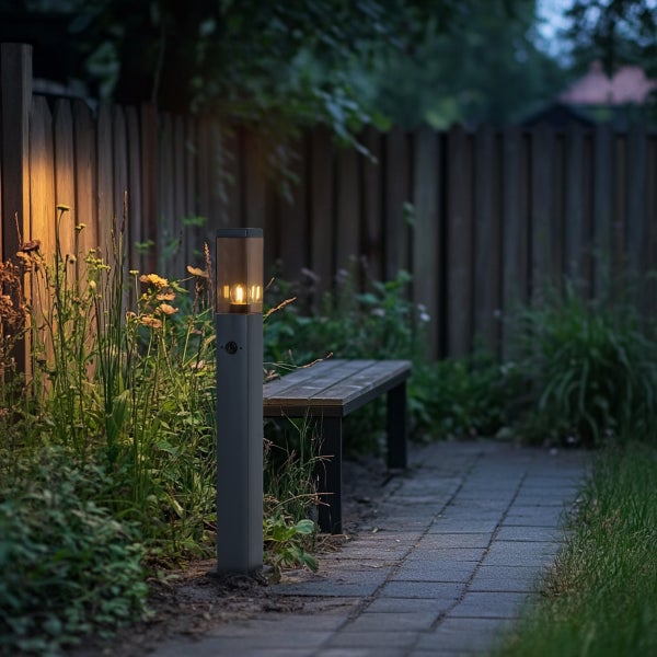 Moderne Außenstehleuchte mit warmer Beleuchtung neben einem Gartenweg aus Stein und einer Holzbank vor einem Holzzaun.