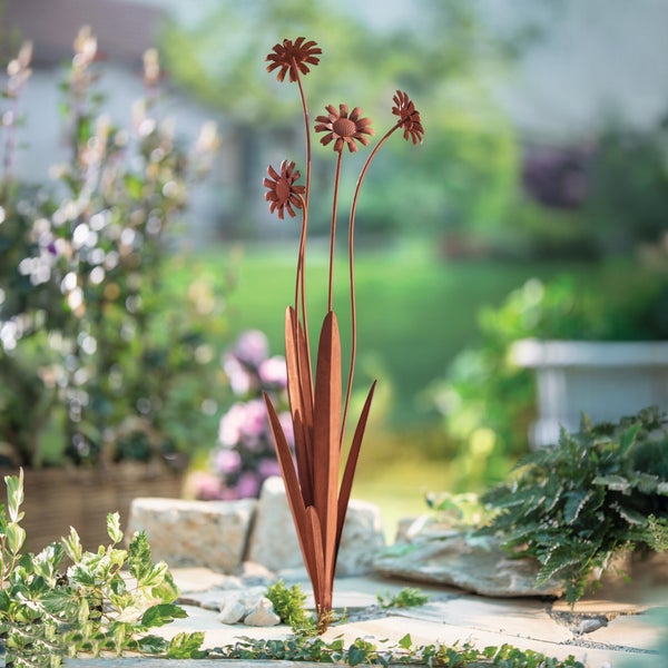 Gartenstecker aus Metall in Blumenform mit Edelrost-Optik in einem hellen Garten zwischen Steinen und Pflanzen.