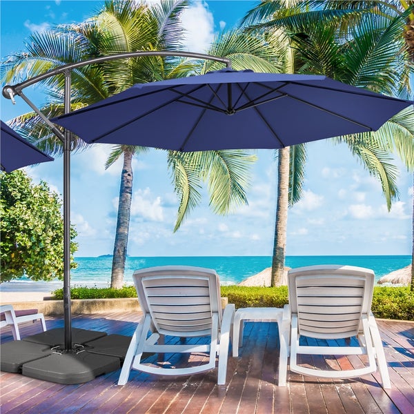 Blauer Ampelschirm mit dunklem Gestell auf einer Holzterrasse mit zwei weißen Sonnenliegen in einer hellen Strandumgebung mit Palmen und Meer.