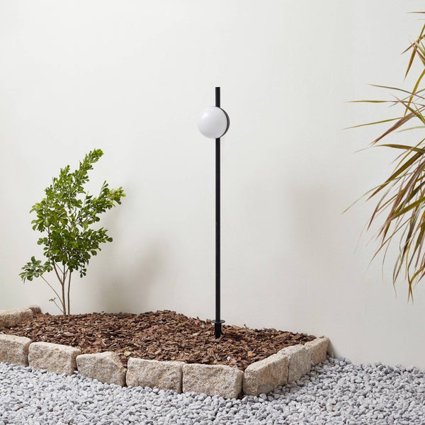 Moderne Außenleuchte mit Kugelschirm auf schwarzem Stab in einem Gartenbeet mit Mulch und Natursteinen.