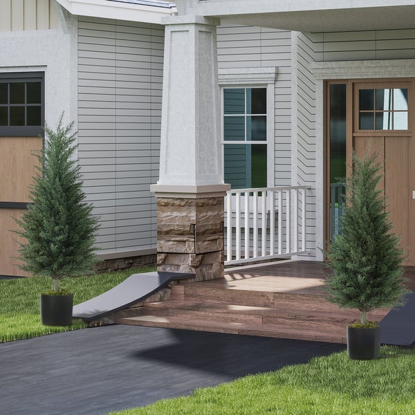 Zwei künstliche Zypressen in schwarzen Töpfen stehen symmetrisch am Hauseingang mit Holzstufen und Steinsäulen.