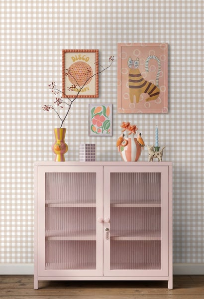 Wohnszene mit beige-weiß karierter Vliestapete, einem rosa Metallschrank mit Riffelglas und dekorativer Wandgestaltung mit gerahmten Bildern.