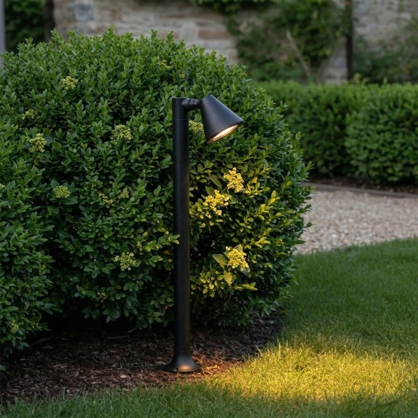 Schwarze Wegeleuchte aus Metall im Garten neben einem Strauch beleuchtet den Rasen mit warmem Licht.