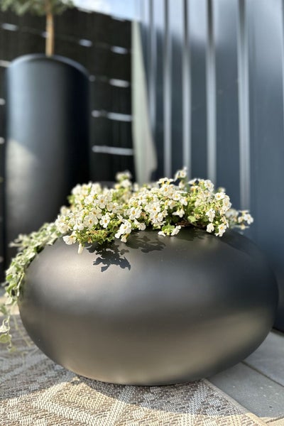 Runder schwarzer Pflanzkübel mit weiß blühenden Blumen auf einer Terrasse.