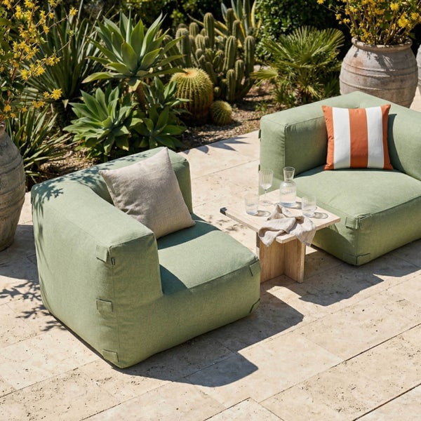 Zwei grüne Outdoor-Lounge-Sessel mit Kissen und ein kleiner Holztisch auf einer hellen Steinterrasse an einem Pool in einem hellen Garten.