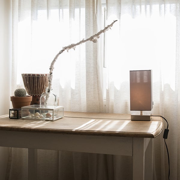 Ein heller Holztisch steht vor einem Fenster mit Vorhängen, dekoriert mit einer Tischlampe, einer kleinen Trommel, Kakteen und Muscheln.