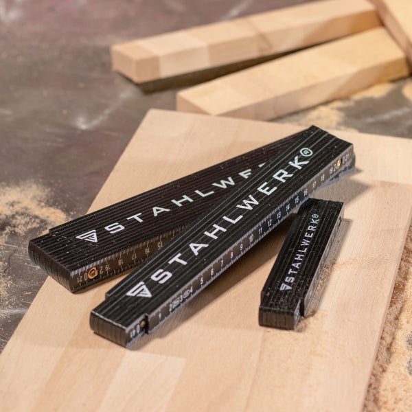 Drei schwarze Gliedermaßstäbe mit STAHLWERK Logo in verschiedenen Größen auf einem hellen Holzbrett mit Sägespänen.